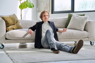 Oturma odasındaki kanepede oturan yakışıklı, rahat bir gencin portresi, kameraya bakan ciddi bir genç. Gençlik yaşı, 16-17 yaş arası erkekler, yaşam tarzı, eğlence konsepti.