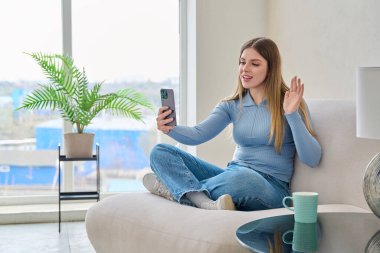 Genç, güzel, gülümseyen bir kadın evinde oturmuş akıllı telefon kullanıyor, mesaj yazıyor ve görüntülü konuşma konferansı veriyor. Teknoloji mobil uygulamaları boş zaman iletişim alışverişi çalışma blogu vlog