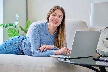 Evdeki kanepede dizüstü bilgisayarla kameraya bakan genç bir kadının portresi. İletişim eğitimi için bilgisayar kullanan güzel bir kadın alışveriş eğitimi iş teknolojisi gençlik yaşam tarzı