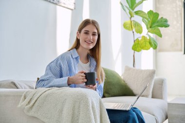 Dizüstü bilgisayarlı, bir fincan kahve içen, evdeki kanepede oturup kameraya bakan güzel, gülümseyen sarışın kadının portresi. Gençlik, yaşam tarzı konsepti