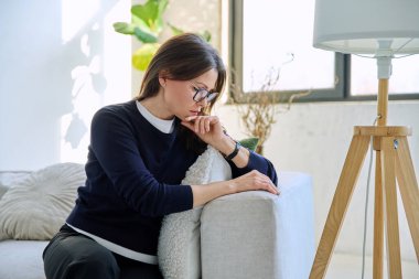 Olgun, üzgün, stresli bir kadın. Stresli, zihinsel ve fiziksel bir acı yaşıyor. Kanepede oturuyor. Sorunlar yalnızlık, baş ağrısı hormon stresi, orta yaşlı kadınların sağlığı