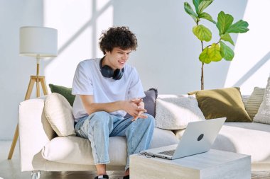 Evdeki kanepede oturmuş dizüstü bilgisayara bakan yakışıklı adam. Boş zamanlarında çalışma iletişim teknolojileri, yaşam tarzı, gençlik kavramı