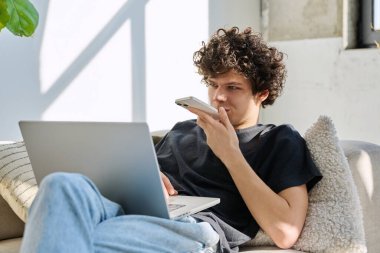 Evdeki kanepede oturan akıllı telefon kullanan, dizüstü bilgisayarı olan yakışıklı, serbest çalışan bir genç. Kıvırcık saçlı ciddi bir adam, modern dijital aletlerle iletişim işi eğitimi alıyor.