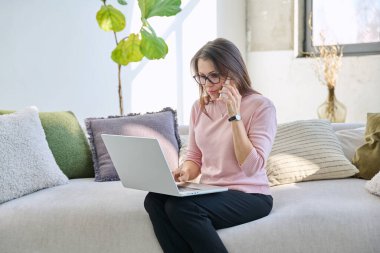 Orta yaşlı bir kadın oturma odasının içinde dizüstü bilgisayarlı akıllı telefonuyla kanepede oturuyor. Olgun kadın iletişim işi için modern dijital cihazlar kullanıyor.