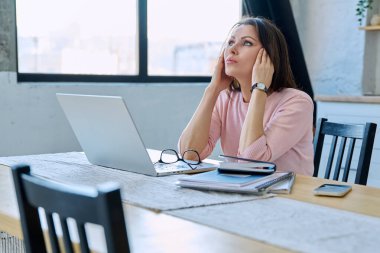 Depresyonda olan, stresli, zihinsel ve fiziksel acı çeken olgun bir kadın bilgisayar laptopuyla masada oturuyor. Sorunlar yalnızlık, baş ağrısı hormon stresi, orta yaşlı kadınların sağlığı