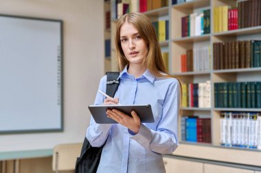 17-18 yaşlarında ciddi bir genç kızın portresi. Sınıfın kütüphanesinde dijital tablet kullanıyor. Gözlüklü sarışın genç bir kadın kameraya bakıyor, eğitim kitaplarıyla dolu raflar.