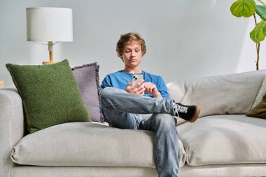 Evdeki kanepede oturan akıllı telefon kullanan yakışıklı, şaşırmış bir genç. Gençlik yaşı, ergenlik, yaşam tarzı, teknoloji konsepti