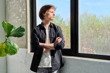 Üzgün, mutsuz, stresli, kolları çaprazlanmış, pencereye bakan genç adam. Duygular, yaşam tarzı, ergenlik kavramı.