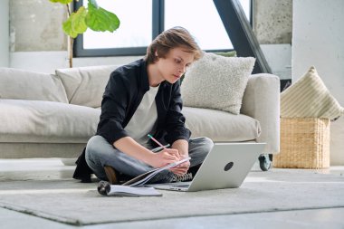 Ciddi bir genç erkek öğrenci dizüstü bilgisayarla evde oturup ders çalışıyor. Eğitim, teknoloji, ergenlik kavramı