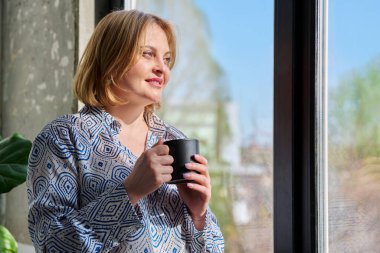 Elinde bardakla pencereden dışarı bakan orta yaşlı güzel bir kadının portresi. Gülümseyen, sarışın, sakin 40 'lı yaşlarda bir kadın. Güzellik, sağlık, yaşam tarzı konsepti