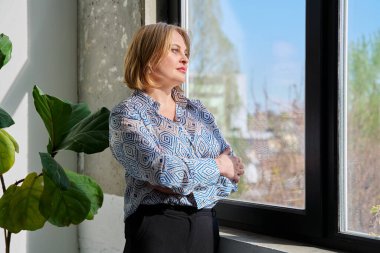 Pencereden dışarı bakan çapraz kollu, kendinden emin, orta yaşlı güzel bir kadının portresi. Gülümseyen, sarışın, sakin 40 'lı yaşlarda bir kadın. Güzellik, sağlık, yaşam tarzı konsepti