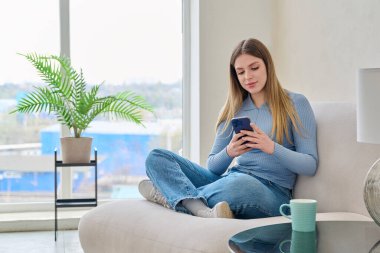 Genç, güzel, gülümseyen bir kadın evinde oturmuş akıllı telefon kullanıyor, mesajlaşıyor, kitap okuyor. Teknoloji mobil uygulamaları serbest iletişim alışverişi çalışma blog gençlik konsepti