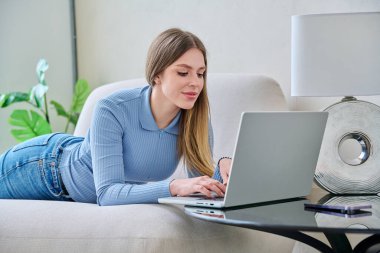 Evde dizüstü bilgisayar kullanarak kanepede yatan genç bir kadın. Gevşemiş güzel bir kadın bilgisayar kullanarak iletişim eğitimi alışveriş işi, oturma odası, teknoloji yaşam tarzı gençliği