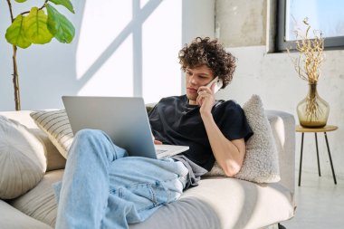 Evdeki kanepede oturan akıllı telefon kullanan, dizüstü bilgisayarı olan yakışıklı, serbest çalışan bir genç. Kıvırcık saçlı ciddi bir adam, modern dijital aletlerle iletişim işi eğitimi alıyor.