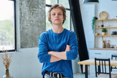 Oturma odasında kollarını kavuşturmuş kendinden emin, ciddi bir gencin portresi. Yakışıklı sarışın adam kameraya bakıyor. Gençlik yaşı, 16-17 yaş arası, ergen, yaşam tarzı, eğlence konsepti