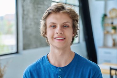 Evdeki oturma odasında kameraya bakan gülümseyen yakışıklı gencin portresi. Gençlik yaşı, 16-17 yaş arası erkekler, ergenlik, eğitim, yaşam tarzı, eğlence kavramı