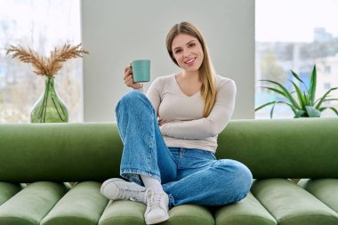 Evdeki kanepede elinde fincanla oturan rahat genç bir kadının portresi. Gülümseyen çekici bir kadın oturma odasında kupayla kanepede dinleniyor. Boş zaman mutluluğu rahat yaşam tarzı gençlik konsepti