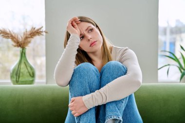 Evdeki kanepede oturan mutsuz, üzgün genç bir kadın, yüzü yakın çekim. Duygusal fiziksel acı çeken üzgün bir kadın. Sorunlar ruhun ve bedenin sağlığı, gençlik kavramı