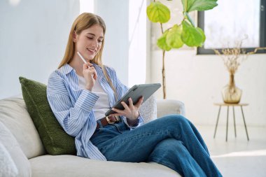 Evdeki kanepede dijital tablet kullanan genç bir kadın. Cihazlı kadın üniversite öğrencisi, rahatlama eğitimi, eğlence iletişimi eğitimi teknolojisi, yaşam tarzı gençlik konsepti