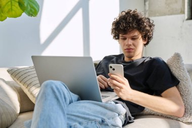 Evdeki kanepede oturan akıllı telefon kullanan, dizüstü bilgisayarı olan yakışıklı, serbest çalışan bir genç. Kıvırcık saçlı ciddi bir adam, modern dijital aletlerle iletişim işi eğitimi alıyor.