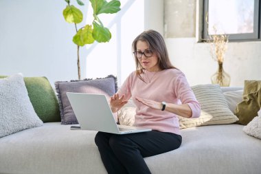 Orta yaşlı bir kadın uzaktan çalışıyor, görüntülü konuşma yapıyor, koltukta dizüstü bilgisayar kullanıyor. Psikoloji oturumu çevrimiçi öğretim koçluğu vlog işi iletişimi