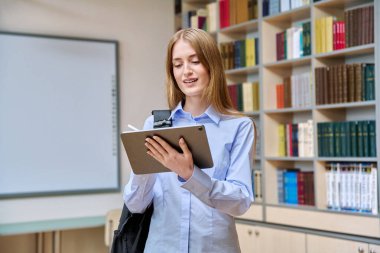 17-18 yaşlarında ciddi bir genç kızın portresi. Sınıfın kütüphanesinde dijital tablet kullanıyor. Gözlüklü sarışın genç kadın, eğitim kitaplarıyla dolu raflar.