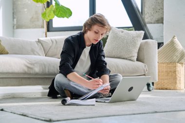 Ciddi bir genç erkek öğrenci dizüstü bilgisayarla evde oturup ders çalışıyor. Eğitim, teknoloji, ergenlik kavramı