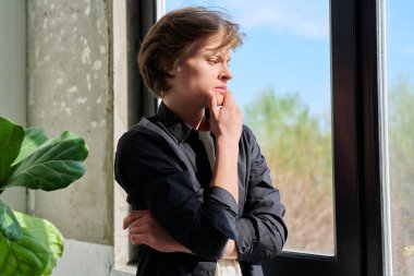 Üzgün, mutsuz, stresli, kolları çaprazlanmış, pencereye bakan genç adam. Duygular, yaşam tarzı, ergenlik kavramı.