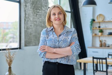 Ev içinde kollarını kavuşturmuş, kendinden emin, orta yaşlı güzel bir kadının portresi. Sarı saçlı, mavi gözlü, kameraya bakan 40 'larında gülümseyen bir kadın. Güzellik, sağlık, yaşam tarzı konsepti