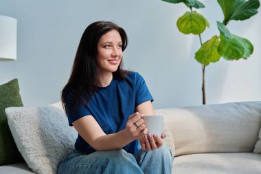 Oturma odasındaki kanepede oturan güzel, gülümseyen esmer kadının portresi. Mutlu, rahatlatıcı, çekici 30 'lu yaşlarda bir kadın başka tarafa bakıyor, kopyalama alanı. Güzellik, yaşam tarzı, kadın konsepti