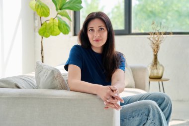 Oturma odasındaki kanepede oturan güzel, mutlu esmer kadının portresi. 30 'lu yaşlarda çekici bir kadın kameraya bakıyor. Güzellik, yaşam tarzı, kadın konsepti