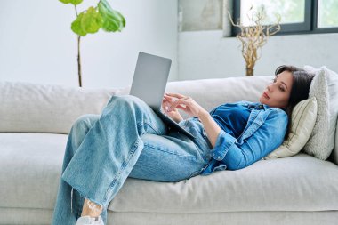 Evdeki kanepede dizüstü bilgisayarda yazan genç ve ciddi bir kadın. Bilgisayarı iş, eğlence, iletişim, alışveriş, serbest yazarlık, blog, eğitim için kullanan bir kadın. Teknoloji yaşam tarzı insan kavramı