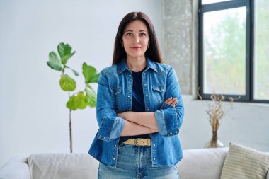 Evin içinde kendine güvenen güzel genç bir kadının portresi. Kollarını kavuşturup kameraya bakan kot elbiseli, gülümseyen kadın. Güzellik, yaşam tarzı, sağlık, 30 'ların insan kavramı