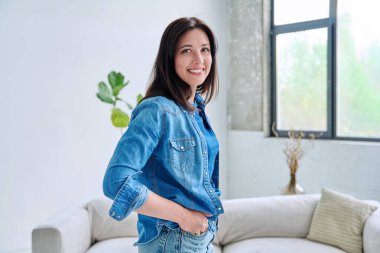 Evdeki güzel genç bir kadının portresi. Kot elbiseli gülümseyen kadın kameraya bakıyor. Güzellik, yaşam tarzı, sağlık, 30 'ların insan kavramı