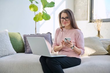Orta yaşlı bir kadın dizüstü bilgisayarlı akıllı telefonuyla kanepede oturuyor oturma odasının içindeki kameraya bakıyor. Olgun gülümseyen kadın iletişim işi için modern dijital cihazlar kullanıyor.