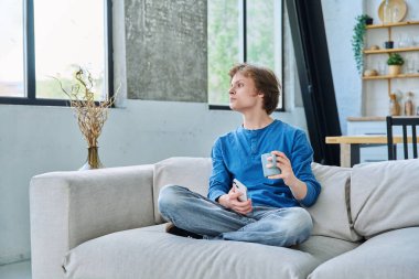 Evde kanepede oturan rahat genç adam elinde akıllı telefon ve bir fincan çayla. Dinlenme, rahatlama, rahatlama, ev hayatı, yaşam tarzı, ergenlik kavramı.