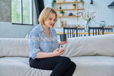 Oturma odasındaki koltukta oturan, elinde akıllı telefon fincanı çayıyla orta yaşlı sarışın kadının portresi. Evin içinde 40 yaşında güzel bir bayan var. Güzellik, sağlık, yaşam tarzı konsepti