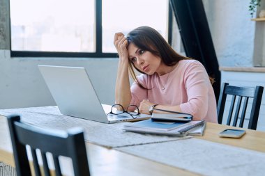 Depresyonda olan, stresli, zihinsel ve fiziksel acı çeken olgun bir kadın bilgisayar laptopuyla masada oturuyor. Sorunlar yalnızlık, baş ağrısı hormon stresi, orta yaşlı kadınların sağlığı
