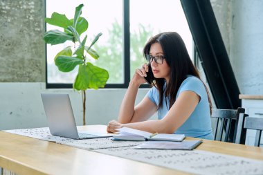 Bilgisayarlı dizüstü bilgisayarla çalışan, akıllı telefondan konuşan, mutfaktaki masada oturan genç bir kadın. Teknoloji, iş, iş, 30 'ların insan kavramı