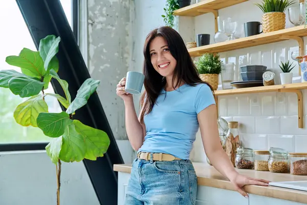 Mutfakta elinde bir fincan kahveyle gülümseyen 30 'lu yaşlardaki güzel bir kadının portresi, kameraya mutlu bir şekilde bakıyor. Yaşam tarzı, ev hayatı, insan kavramı.