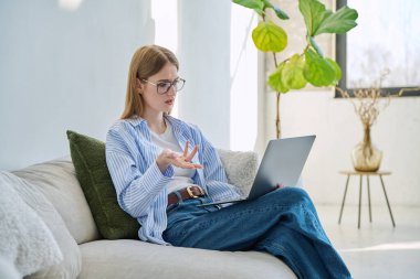 Genç kız video sohbet konferansı için dizüstü bilgisayar kullanıyor. Kanepede oturmuş web kamerasına bakıyor, bilgisayar ekranından konuşuyor. Eğitim eğitimi teknolojileri gençlik iletişimi kavramı üzerinde çalışır