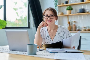 Orta yaşlı gülümseyen güzel bir kadın dizüstü bilgisayar belgeleriyle çalışıyor, mutfaktaki masada oturan kameraya bakıyor. Teknoloji, iş, iş, olgun insanlar konsepti