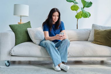 Ciddi bir genç kadın akıllı telefon kullanarak kanepede oturuyor. İletişim, eğlence, alışveriş, iş ve eğitim için mobil başvurular. Teknoloji yaşam tarzı insan kavramı