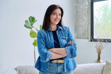 Evin içinde kendine güvenen güzel genç bir kadının portresi. Kollarını kavuşturup kameraya bakan kot elbiseli, gülümseyen kadın. Güzellik, yaşam tarzı, sağlık, 30 'ların insan kavramı
