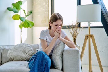 Üzgün, hayal kırıklığına uğramış, 17-18 yaşlarında bir genç kız evinde kanepede oturuyor. Stres, depresyon, gençlik, gençlik krizi, gençlerin akıl sağlığı konsepti