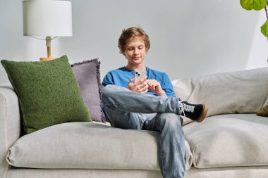Evdeki kanepede oturan akıllı telefon kullanan yakışıklı genç adam. Gençlik yaşı, ergenlik, yaşam tarzı, teknoloji konsepti