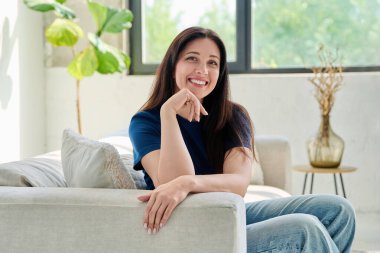 Oturma odasındaki kanepede oturan güzel, mutlu esmer kadının portresi. 30 'lu yaşlarda çekici bir kadın kameraya bakıyor. Güzellik, yaşam tarzı, kadın konsepti