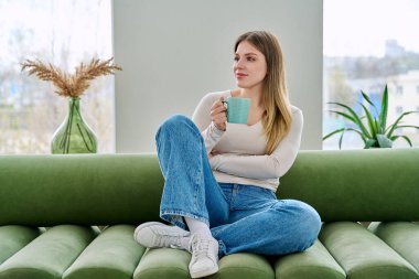 Evdeki kanepede elinde fincanla oturan rahat genç bir kadının portresi. Gülümseyen çekici bir kadın oturma odasında kupayla kanepede dinleniyor. Boş zaman mutluluğu rahat yaşam tarzı gençlik konsepti