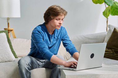 Evde kanepede oturup dizüstü bilgisayarda daktilo eden yakışıklı bir delikanlı. Gençlik yaşı, ergenlik, yaşam tarzı, teknoloji konsepti