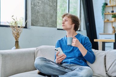 Evde kanepede oturan rahat genç adam elinde akıllı telefon ve bir fincan çayla. Dinlenme, rahatlama, rahatlama, ev hayatı, yaşam tarzı, ergenlik kavramı.